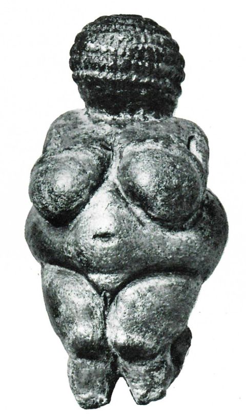 Sculpture. Reproduced in black and white, this statuette has a bulbous, textured orb for a head, bulging breasts and belly, a deep navel, defined pubis, and legs that taper or have been broken.