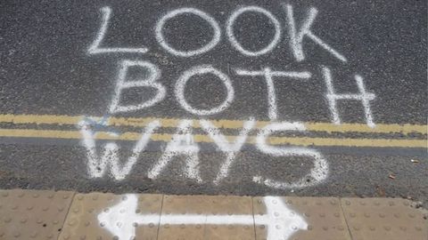 Graffiti art. A street is spray-painted in white with the words 'Look both ways' above a double-sided arrow. Double yellow lines cross the picture, and textured pavers indicate that this is where a sidewalk meets the street.