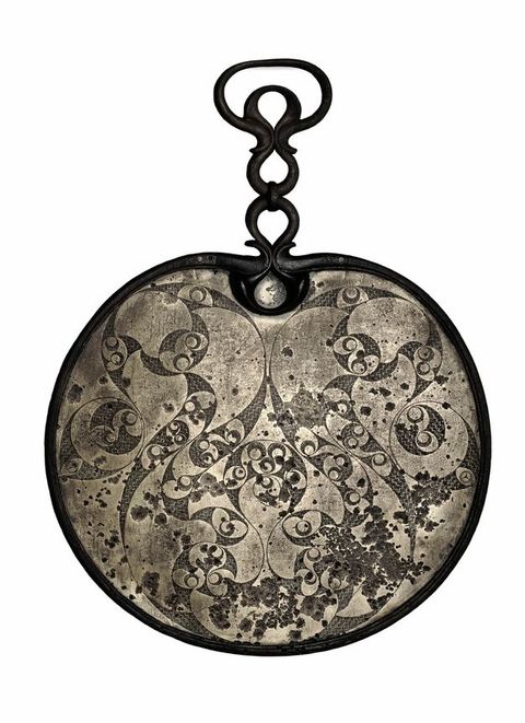 Mirror. The back of this bronze mirror is decorated with dark, swirling leaf-like and circular designs against a brighter background. A handle or hook at the top is made of three teardrop-shaped ornaments, the topmost set inside a larger oval.