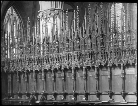 Black and white photograph. Individual seats along a bench-like row of a choir stall are separated by arms that project from a high back. Using the chairs for scale, the screen appears to be one or two stories high, and it is elaborately carved with tracery, spires, and figures.